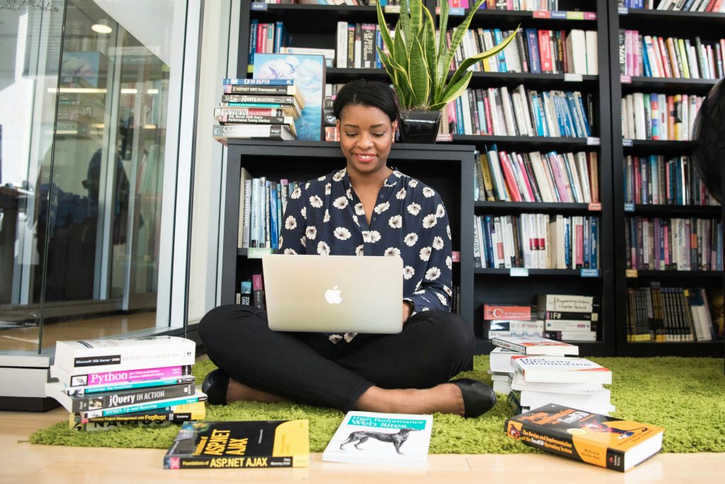 A woman sitting on a carpet in a library working on her laptop, surrounded by books.
